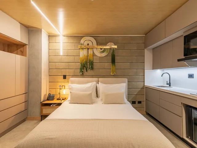 Apartment-style suite at Living by Armóniko with a double bed, upholstered headboard, green and yellow macramé on a block wall, light built-in wardrobe with a wood niche and a kitchenette with sink, cooktop, microwave and minibar