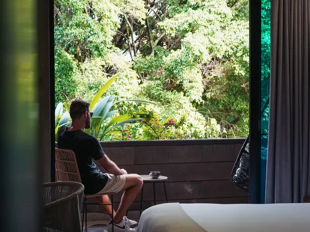 Guest sitting on his suite balcony at Living by Armóniko looking out at leafy trees in Laureles, with a cup of coffee on a side table and the bed in the foreground