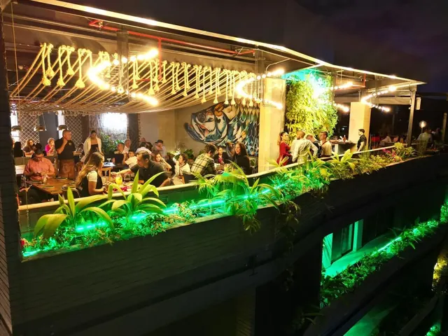 Rocoto Rooftop terrace at night on the fifth floor of Living by Armóniko, with a rope ceiling and warm string lights, a large fish mural, a vertical garden and diners at tables beside illuminated planters