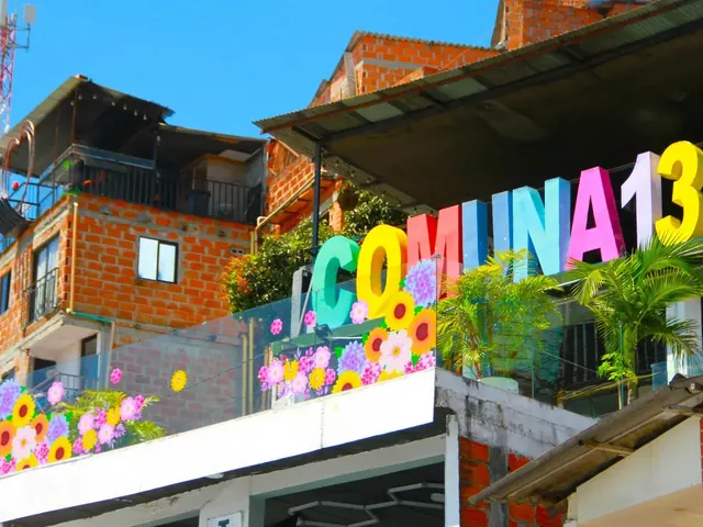 Colorful letters spelling COMUNA 13 on the terrace of a brick house decorated with painted flowers and palms, under a blue sky in Medellín