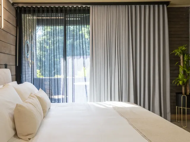Living by Armóniko suite with a white-linen bed and cream pillows, a grey curtain and black sheer in front of the glass balcony door, trees outside and a plant in a black pot on a gold stand