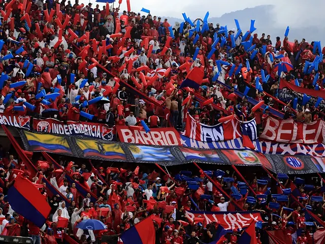 Fans in red and blue shirts, waving red and blue flags and inflatable sticks in Independiente Medellín's colors, pack a stadium stand hung with banners, with cloudy mountains behind