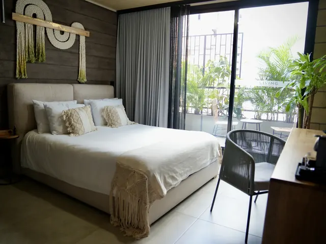 Living by Armóniko suite with a bed in white bedding, macramé pillows and a rope wall hanging, and a glass door with grey curtains opening onto a balcony with plants, a small table and chairs