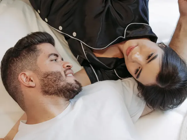 Smiling couple lying on the white bed of a Living by Armóniko suite, she in black satin pajamas and he in a white T-shirt