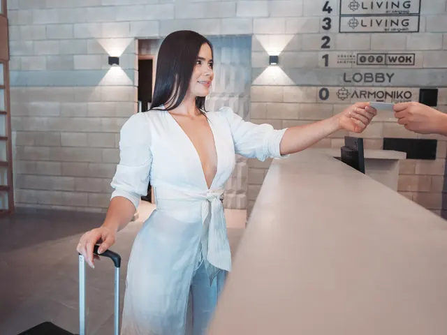 Guest dressed in white with a black suitcase receives a card at the Living by Armóniko front desk, beside the floor directory listing Rocoto and 123 Wok
