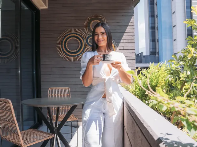 Smiling woman holding a cup of coffee on the balcony of a Living by Armóniko suite, with a table, wicker chairs, plants and tall buildings in the background