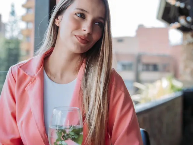 Woman in a pink coat holding a glass with fresh mint at a wooden table on the Living by Armóniko rooftop, under hanging bulbs, with Laureles brick buildings in the background