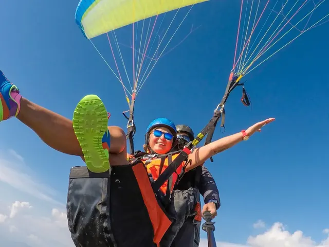 Smiling passenger in a blue helmet and sunglasses flying a tandem paraglider with her pilot under a lime-green wing and blue sky