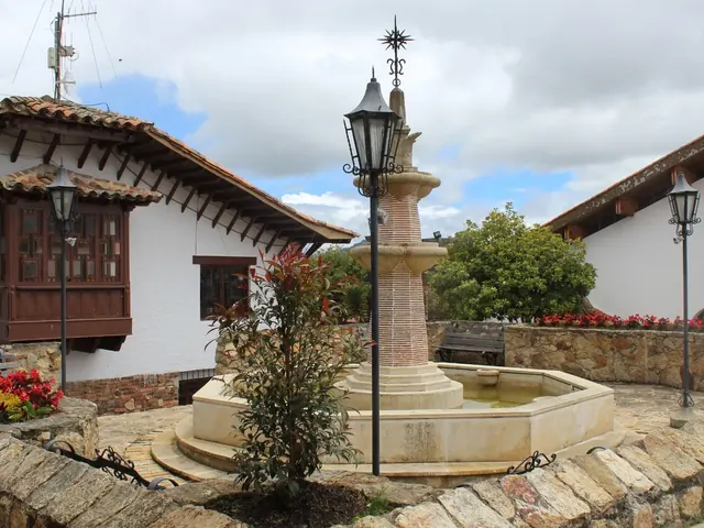 Colonial-style square with a stone fountain, iron lampposts, planters of red flowers and whitewashed houses with clay-tile roofs and a wooden balcony