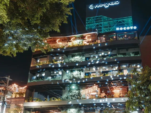 Living by Armóniko building at night, with plant-filled balconies, illuminated Armóniko, Rocoto and Torrealta signs, and a yellow vintage jeep in front of a flower arch at the entrance