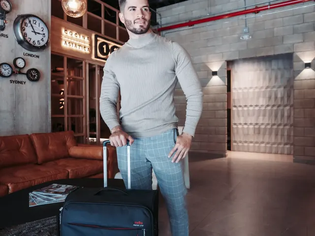 Traveler in a turtleneck sweater with a black suitcase in the Living by Armóniko lobby, with world-city clocks on the wall, a leather sofa and pendant lights