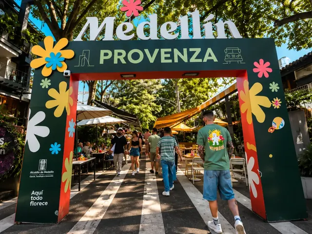 Green welcome arch with colorful flowers and a Medellín Provenza sign over a tree-lined street in El Poblado, with terraces and people walking by day