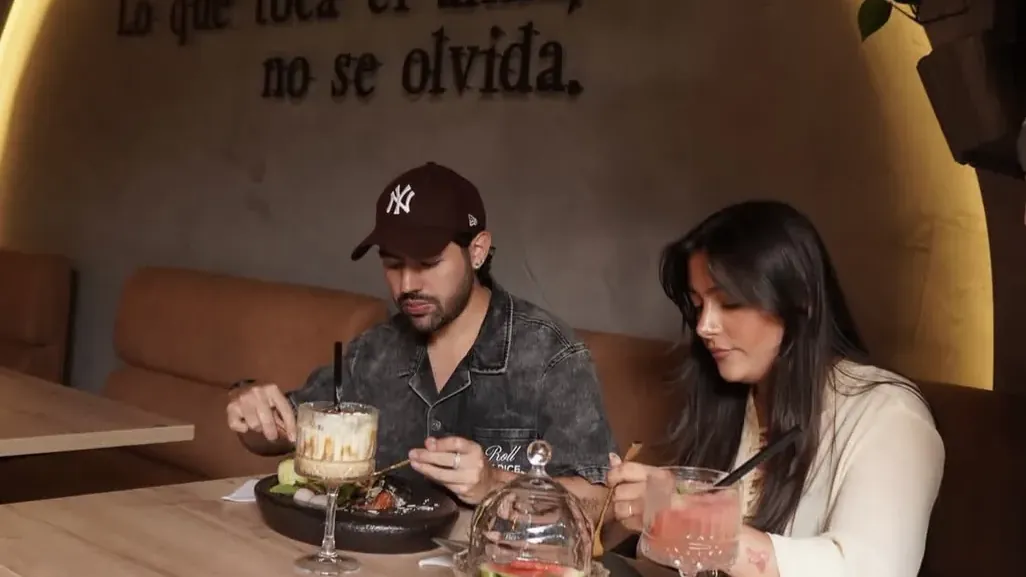 Couple sharing cocktails and dishes at a wooden table beneath a lit arch with the Spanish phrase Lo que toca el alma, no se olvida