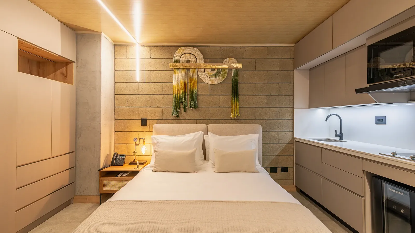 Apartment-style suite at Living by Armóniko with a double bed, upholstered headboard, green and yellow macramé on a block wall, light built-in wardrobe with a wood niche and a kitchenette with sink, cooktop, microwave and minibar