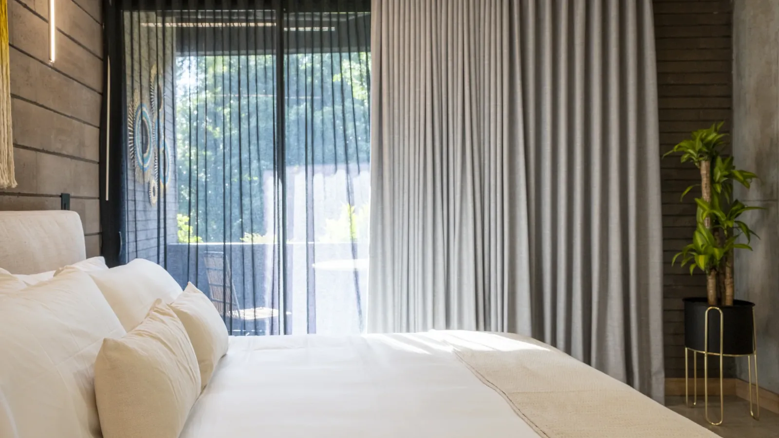 Suite de Living by Armóniko con cama de lencería blanca y cojines crema, cortina gris y visillo negro frente a la puerta de vidrio del balcón, árboles al fondo y una planta en matera negra con base dorada