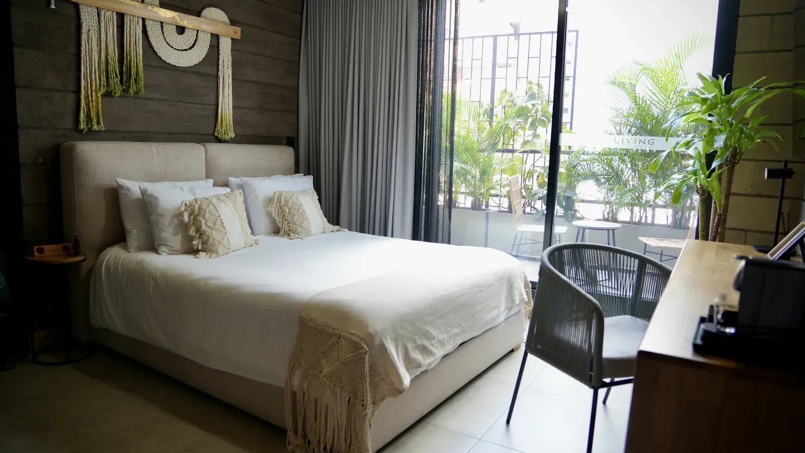 Suite de Living by Armóniko con cama de ropa blanca y cojines de macramé, tapiz de cuerdas en la pared y puerta de vidrio con cortinas grises que abre a un balcón con plantas, mesa y sillas