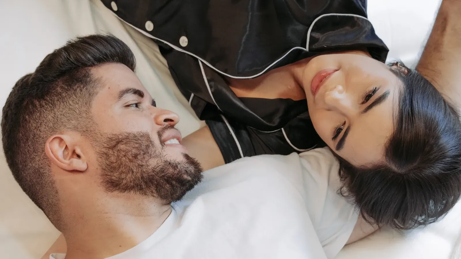 Smiling couple lying on the white bed of a Living by Armóniko suite, she in black satin pajamas and he in a white T-shirt