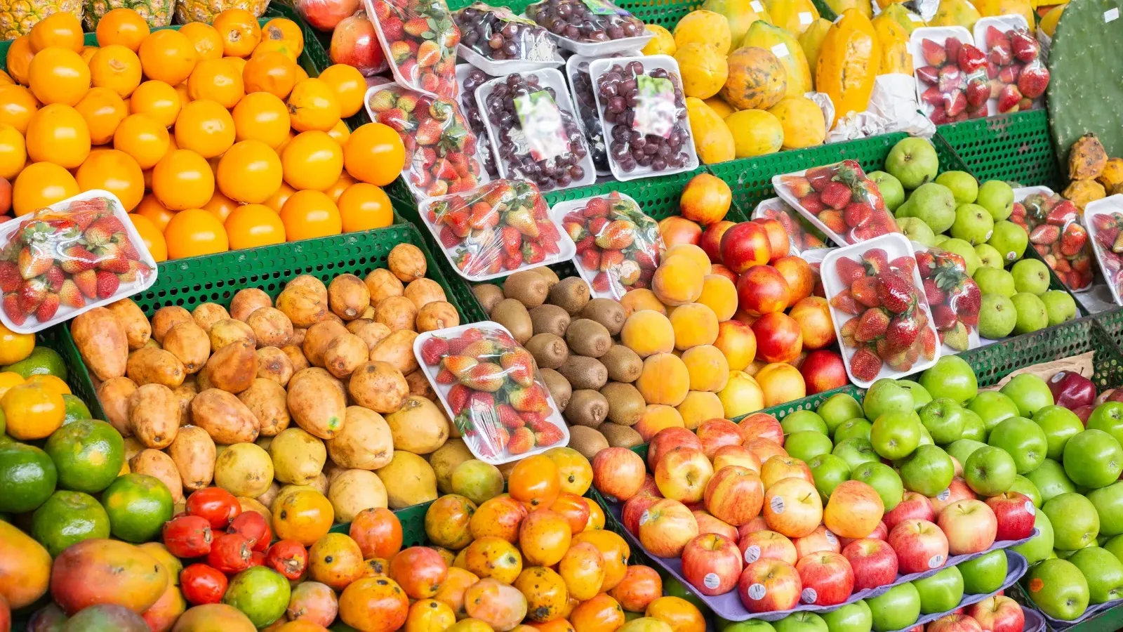 Puesto de frutas en un mercado con naranjas, fresas en bandejas, uvas, papayas, kiwis, manzanas, mangos y pitahayas amarillas en canastas verdes