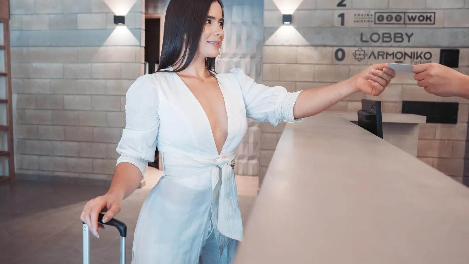 Guest dressed in white with a black suitcase receives a card at the Living by Armóniko front desk, beside the floor directory listing Rocoto and 123 Wok
