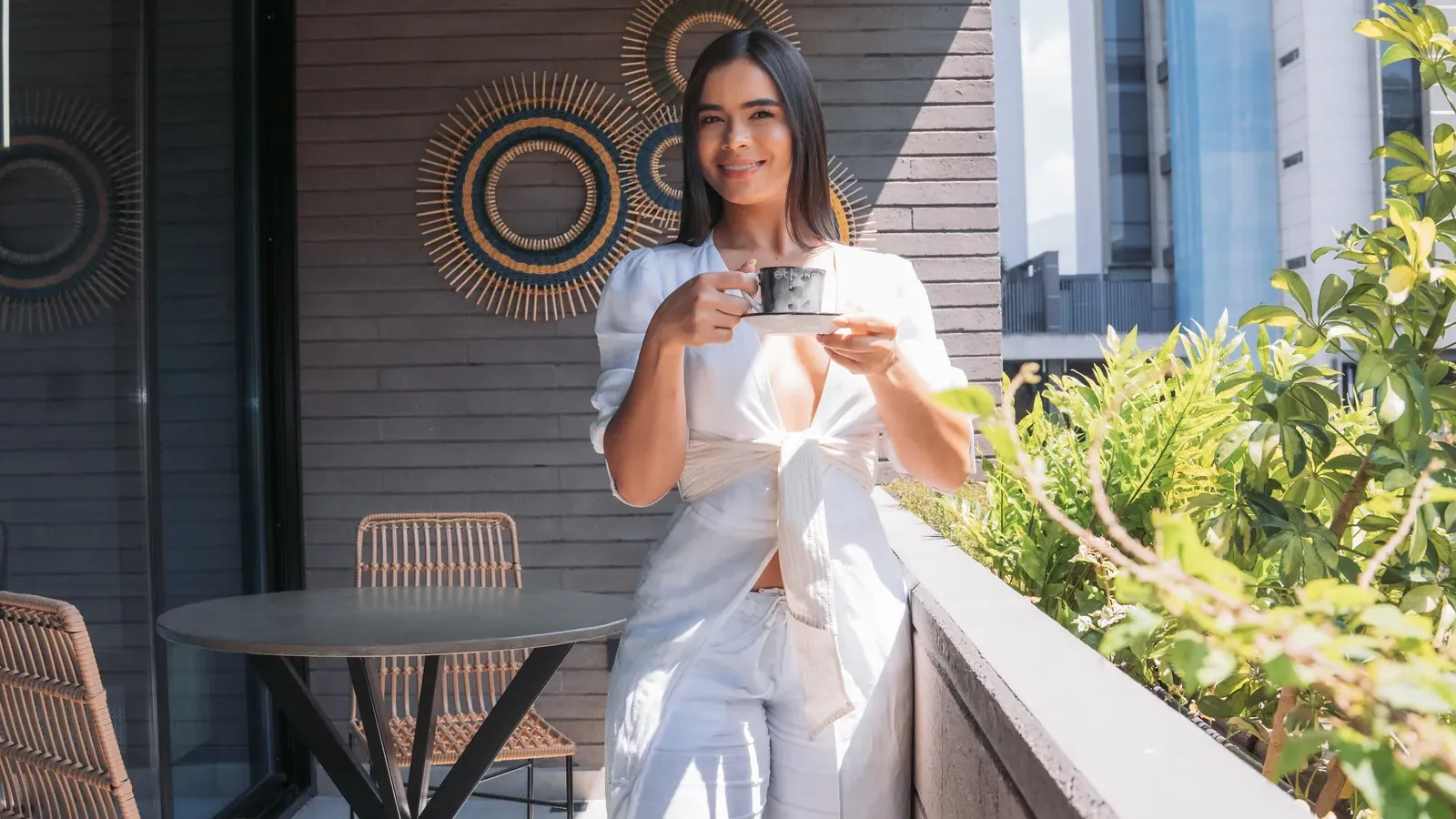 Smiling woman holding a cup of coffee on the balcony of a Living by Armóniko suite, with a table, wicker chairs, plants and tall buildings in the background