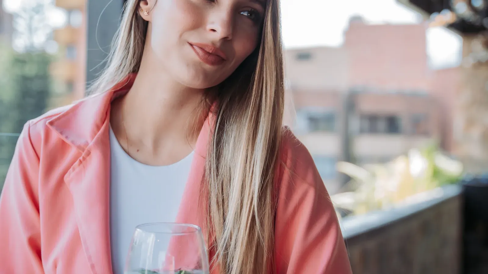 Mujer con abrigo rosado y copa con hierbabuena en una mesa de madera del rooftop de Living by Armóniko, bajo bombillos colgantes y con edificios de ladrillo de Laureles al fondo