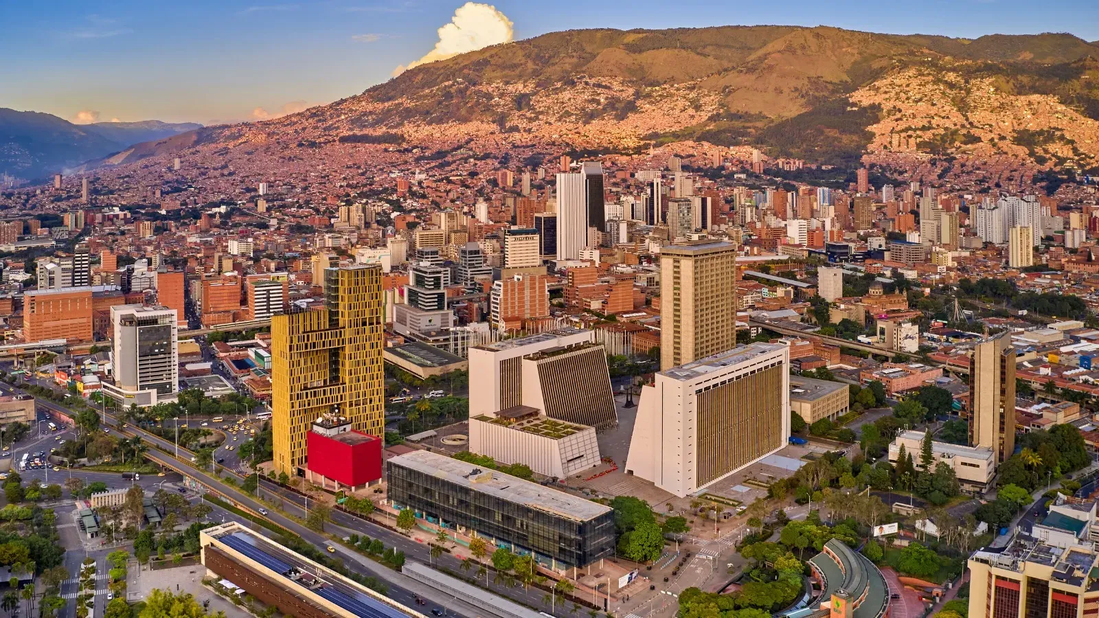 Aerial view of downtown Medellín at sunset, with administrative towers, tree-lined avenues and the Aburrá Valley mountains in the background