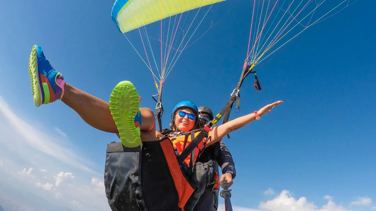 Pasajera sonriente con casco azul y gafas de sol vuela en parapente biplaza junto a su piloto, bajo una vela verde limón y el cielo azul