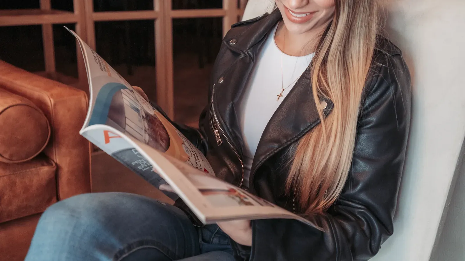 Huésped sonriente leyendo una revista en una silla lounge blanca en el lobby de Living by Armóniko, junto a un sofá de cuero color coñac y con una estantería de madera en retícula al fondo