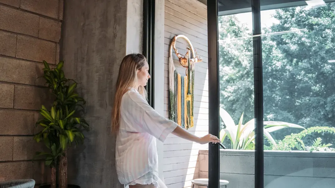 Huésped en pijama abre la puerta corrediza de vidrio de su suite hacia un balcón con plantas y árboles, con la cama en primer plano