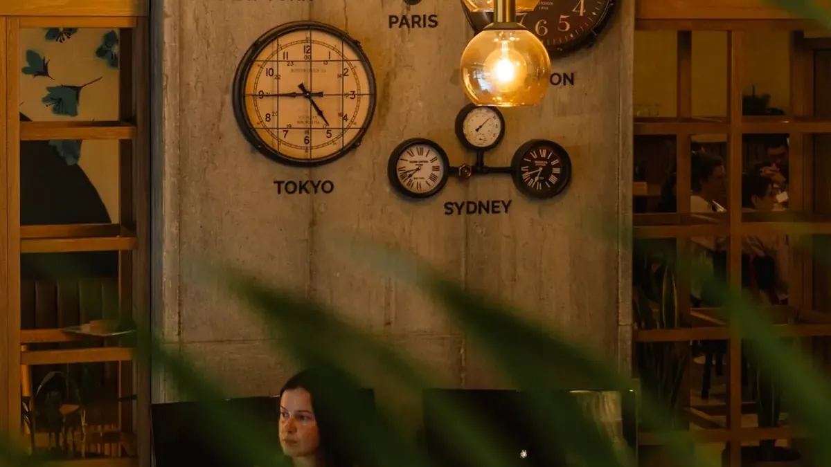 Lobby of Living by Armóniko: a concrete column with clocks showing the time in New York, Paris, Tokyo and Sydney, amber glass pendant lamps and a guest sitting below, seen through palm leaves