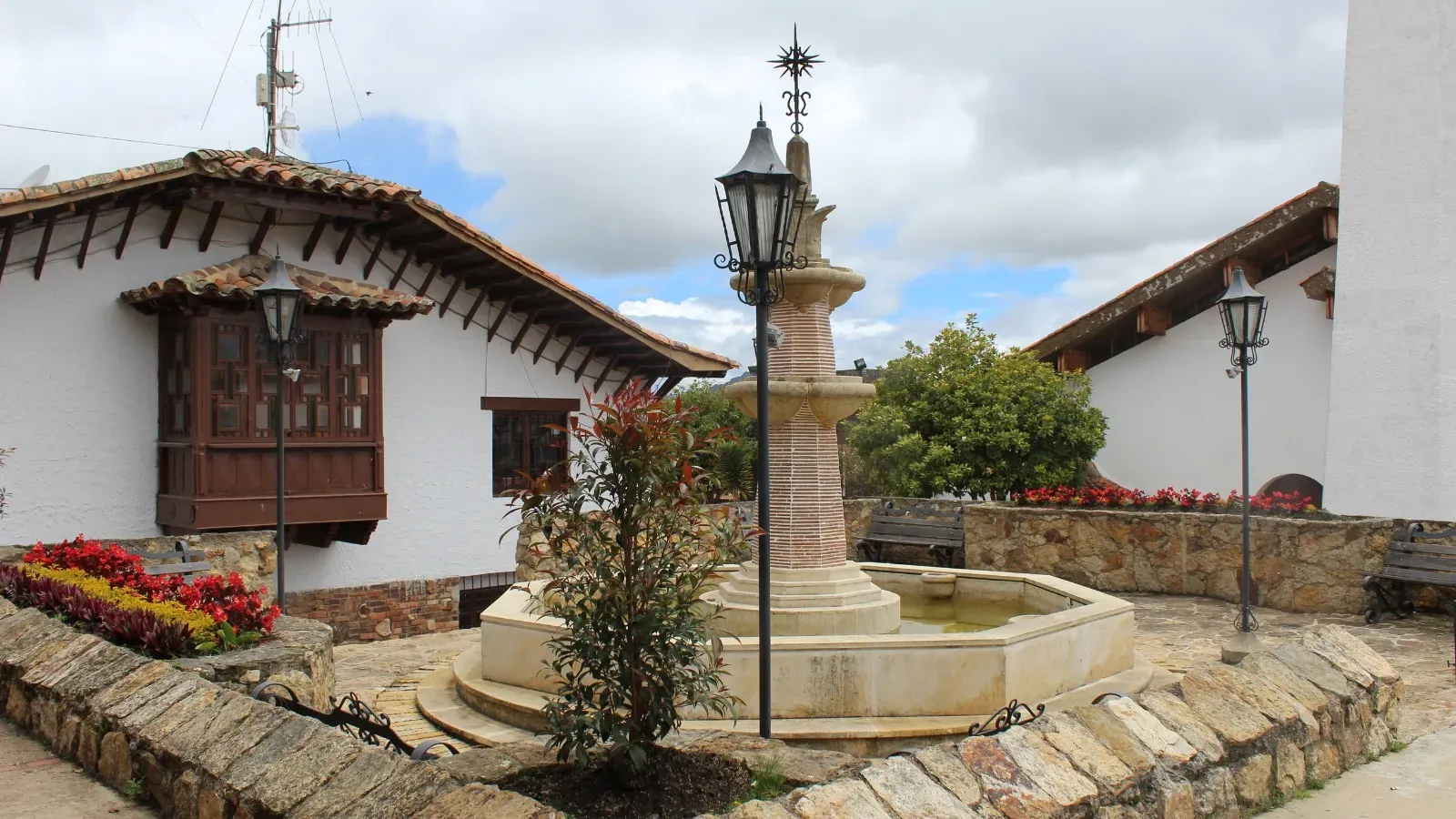 Plazuela de estilo colonial con fuente de piedra, faroles de hierro, jardineras con flores rojas y casas encaladas con tejas de barro y un balcón de madera