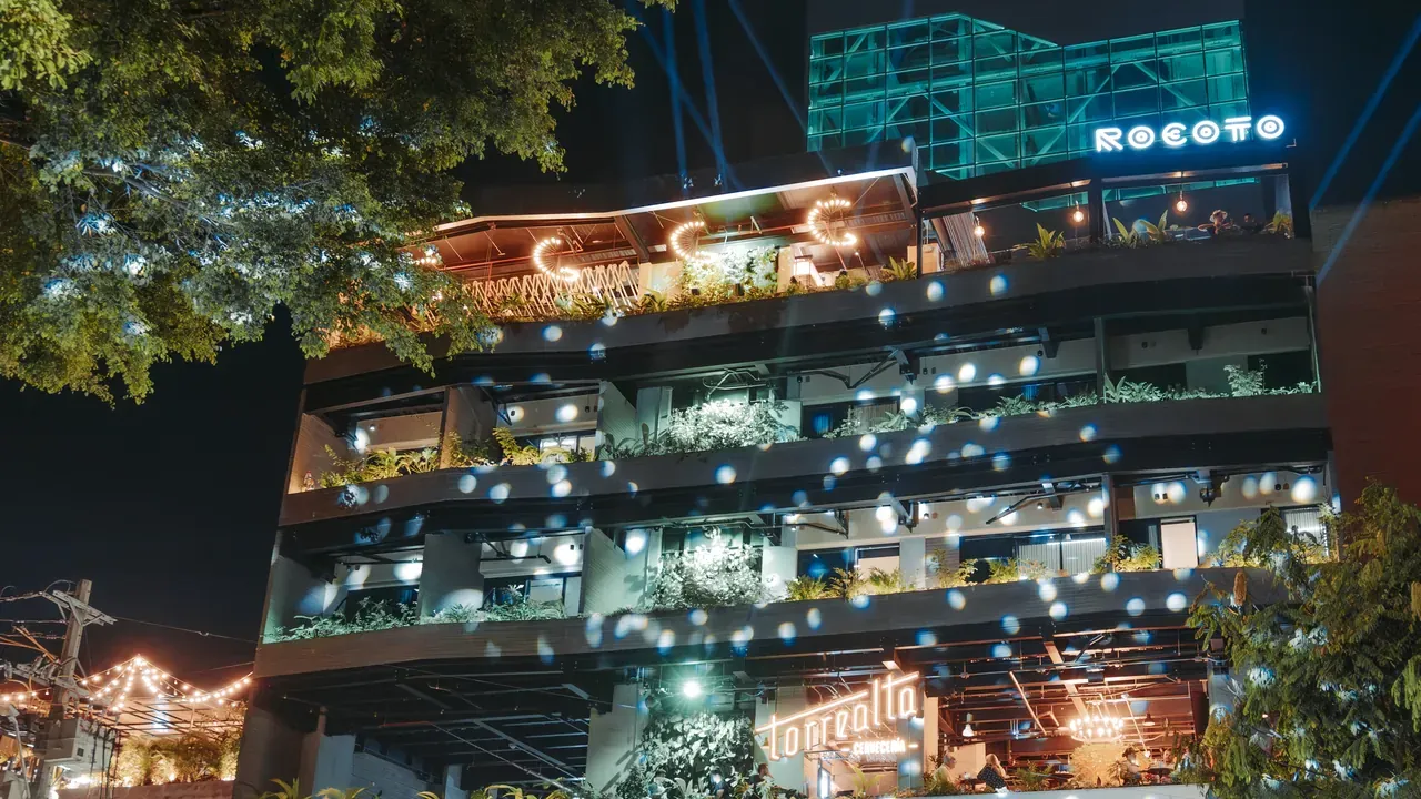 Fachada de Living by Armóniko de noche, con balcones llenos de plantas, los letreros iluminados de Armóniko, Rocoto y Torrealta, y un campero amarillo frente a un arco de flores en la entrada