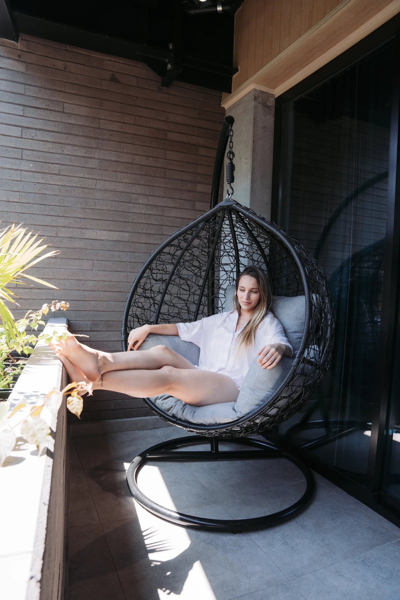 Huésped descansa en una silla colgante sobre un balcón soleado de Living by Armóniko, junto a plantas y una pared de ladrillo