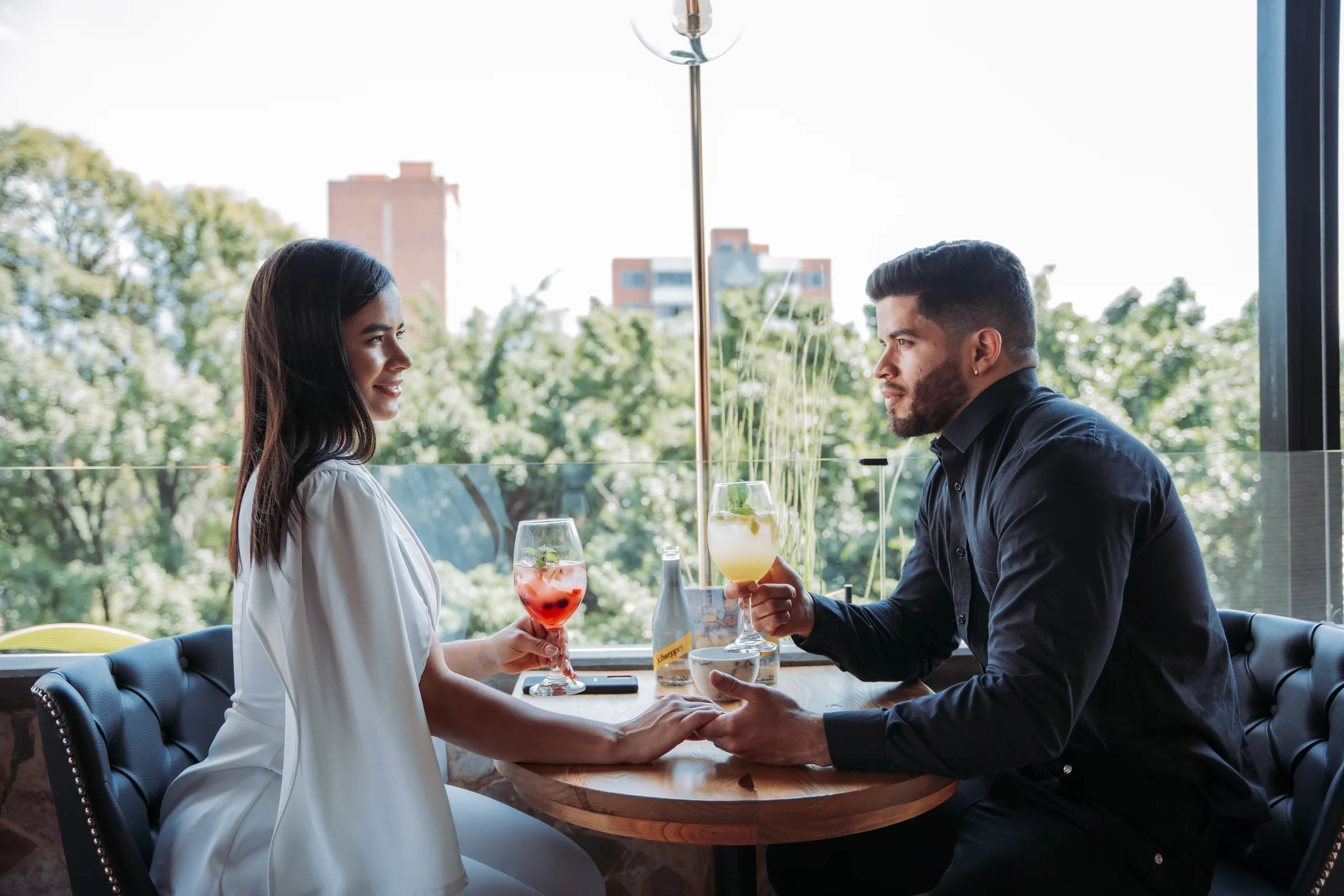 Couple holding hands at a table with cocktails by the window at Living by Armóniko, Laureles
