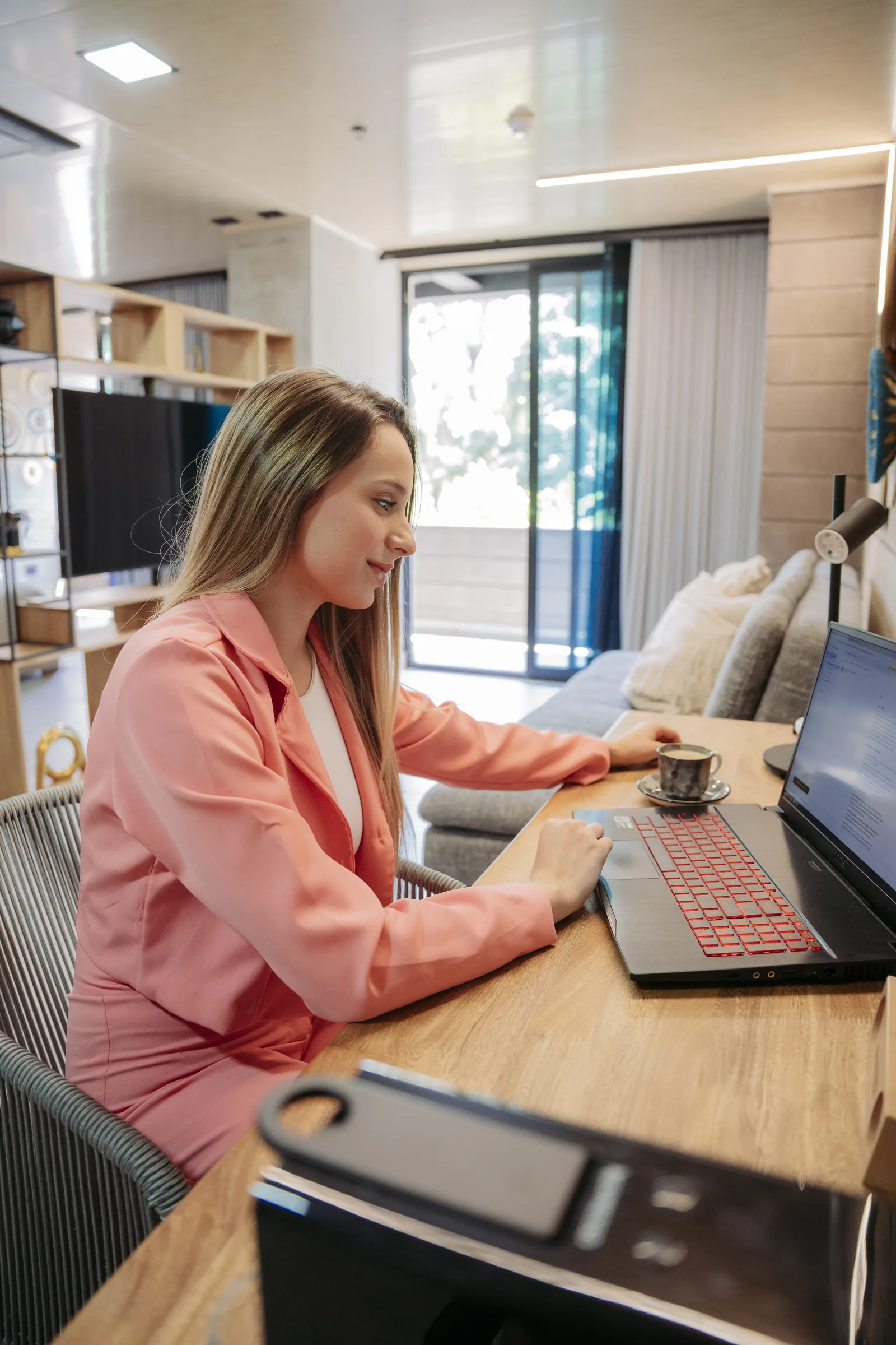 Guest working on a laptop at a wooden desk inside a suite with a sofa, TV and a door to the balcony