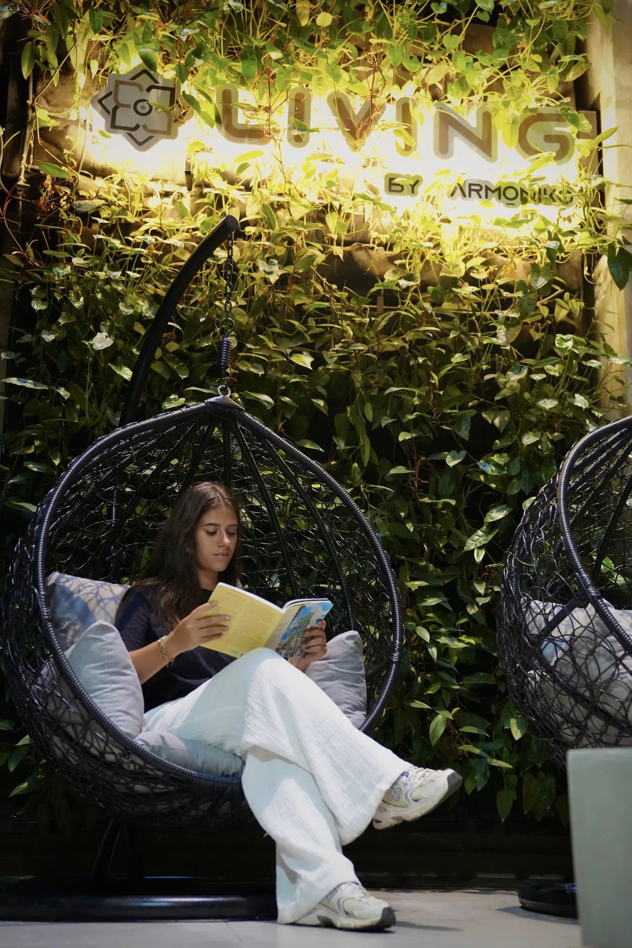 Huésped leyendo un libro en una silla colgante tejida frente a un jardín vertical con el letrero iluminado de Living by Armóniko