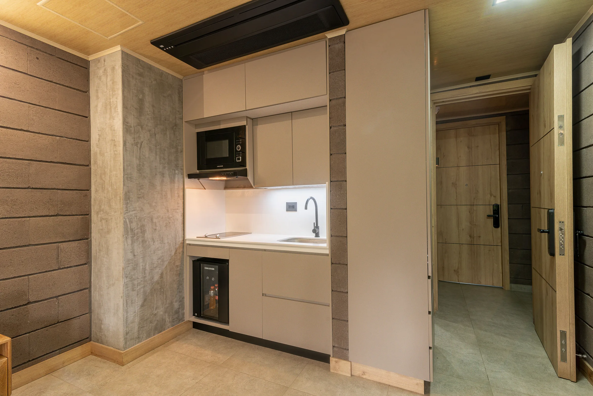 Kitchenette in a Comfort Suite at Living by Armóniko with built-in cooktop, microwave, sink, light cabinets and minibar, beside the entrance hallway