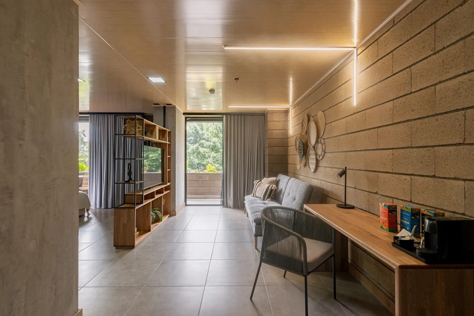 Sala de la Suite Deluxe de Living by Armóniko con escritorio de madera, silla tejida gris, sofá y puerta al balcón con vista a los árboles