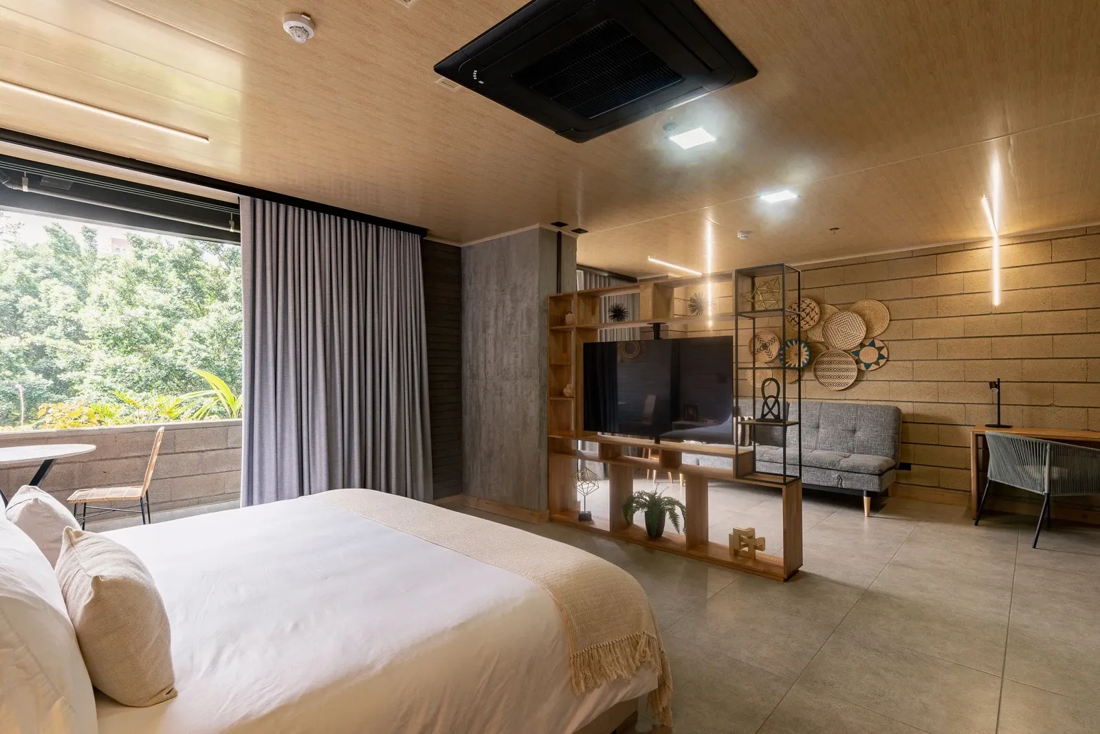 Living by Armóniko Deluxe Suite with a king bed, gray curtains open to a tree-lined balcony, a wooden divider unit with a TV, a gray sofa and a desk