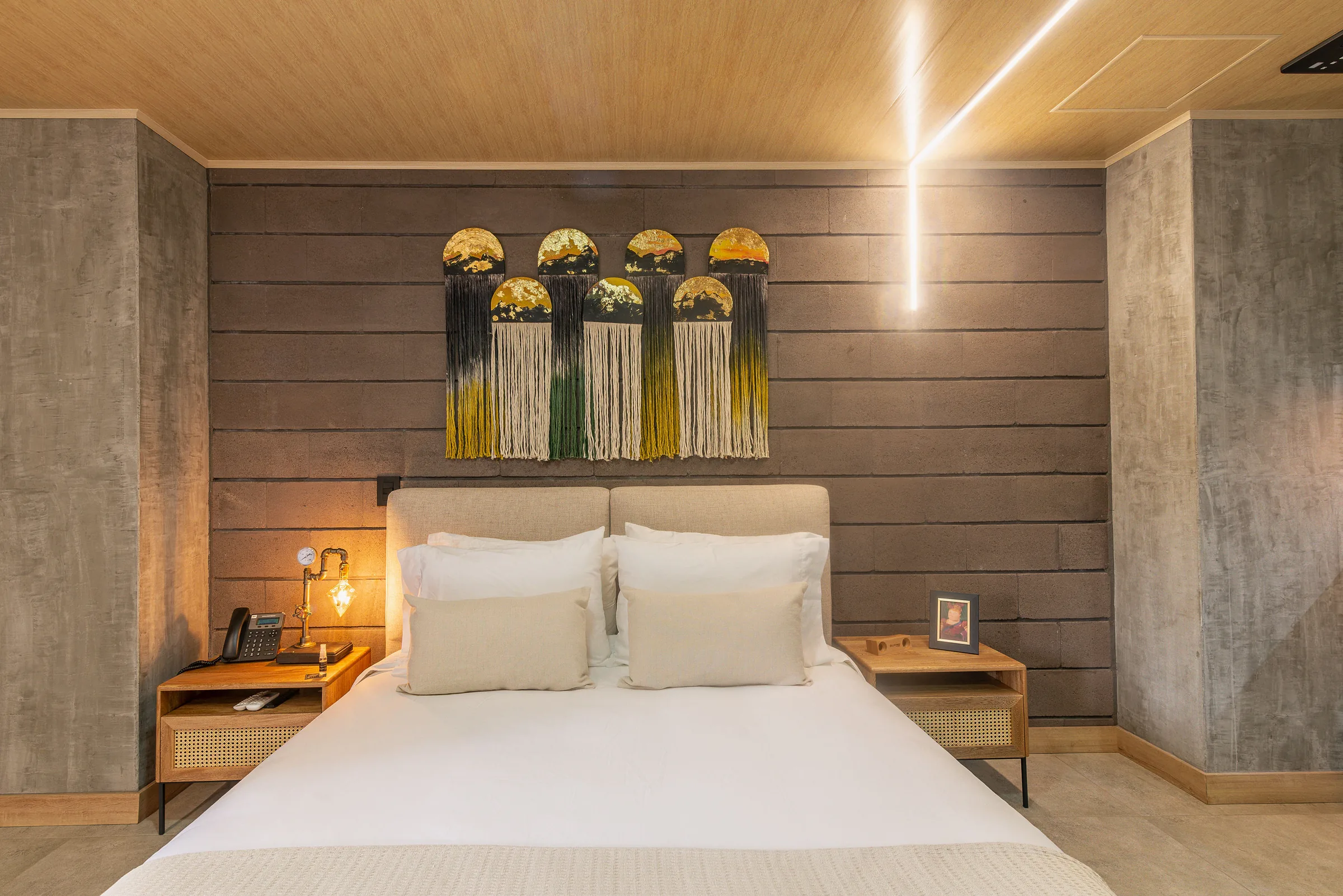 Suite Superior de Living by Armóniko: cama Queen de lencería blanca bajo un tapiz de macramé con medias lunas doradas y flecos de colores, mesas de noche de madera y ratán y una lámpara industrial encendida