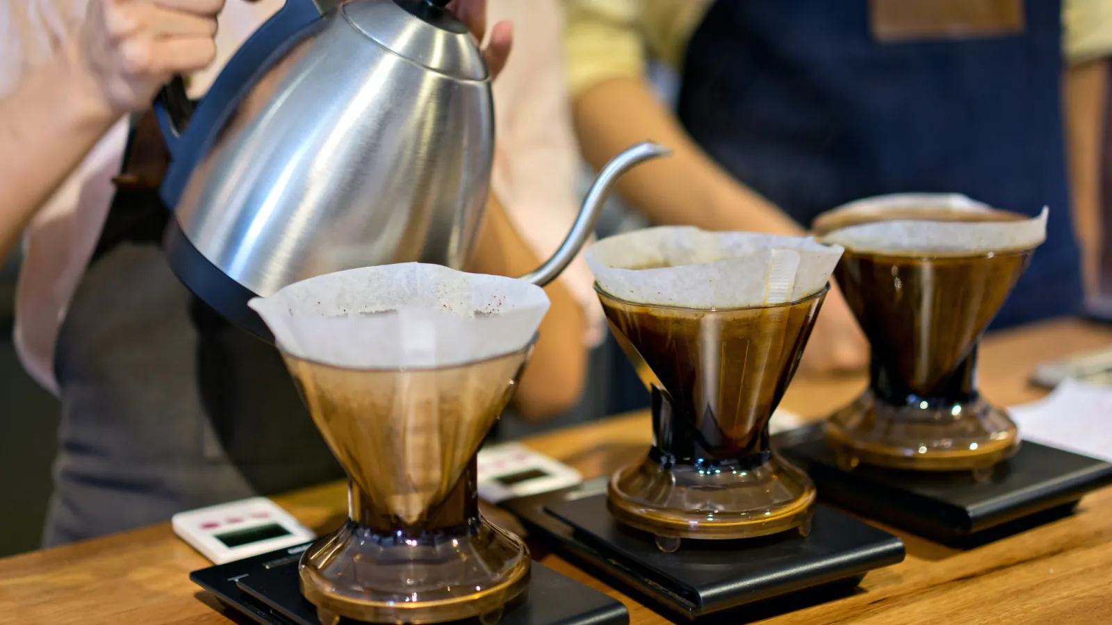 Barista vierte agua caliente con una tetera de cuello de cisne sobre tres goteros con filtro de papel, cada uno apoyado en una báscula digital