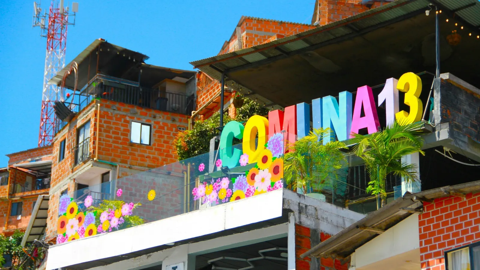 Colorful letters spelling Comuna 13 on a terrace decorated with painted flowers, surrounded by brick houses in Medellín
