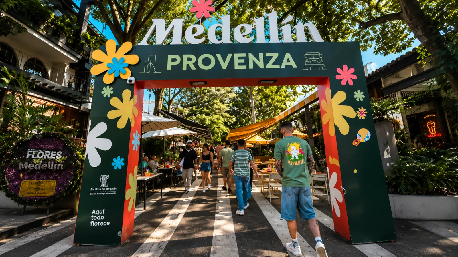 Entrance arch to Provenza in El Poblado with a Medellín Provenza sign decorated with colorful flowers and people walking down a tree-lined street with terraces